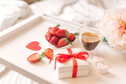 Breakfast for Valentine's Day. Heart shaped white plate with fresh strawberries, cup of coffee, gift and flowers with gift in bed. Still life composition.