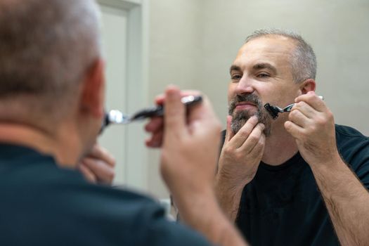 Serious handsome man looking in mirror and shaving his whiskers to look younger