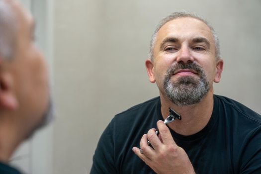 Charming middle aged man shaving his beard and moustache off in front of mirror to look younger. High quality photo