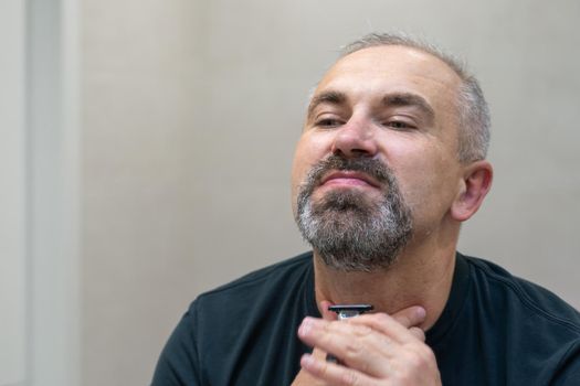 Charming middle aged man shaving his beard and moustache off in front of mirror to look younger. Mature gray-haired man restyling his beard himself at home using razor