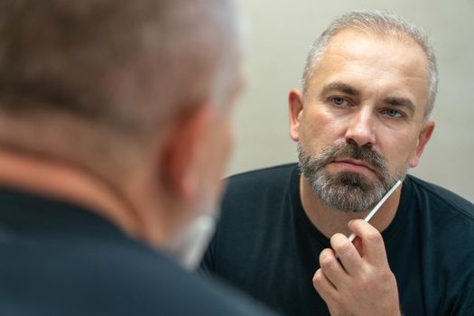 Middle-aged handsome man brushing his beard in the morning in bathroom looking in mirror. Face care concept