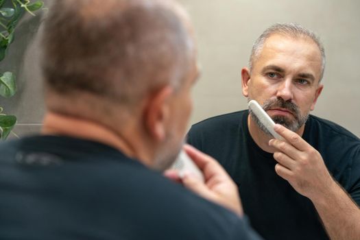 Middle-aged handsome man brushing his beard in the morning in bathroom looking in mirror. Face care concept