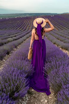 A young beautiful girl in a purple flying dress stands on a blooming lavender field. Rear view. The model has a straw hat.