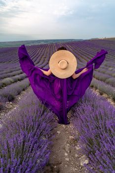 A young beautiful girl in a purple flying dress stands on a blooming lavender field. Rear view. The model has a straw hat.