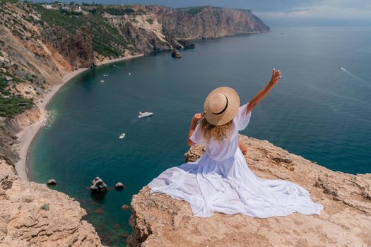 Portrait of a young woman on the beach by the sea, sitting with raised hands in a white dress and hat. Back photo.