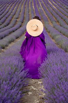 A young beautiful girl in a purple flying dress stands on a blooming lavender field. Rear view. The model has a straw hat.