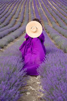 A young beautiful girl in a purple flying dress stands on a blooming lavender field. Rear view. The model has a straw hat.