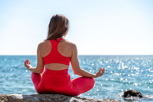 A young woman in red leggings and a red top with long loose hair practices yoga outdoors by the sea on a sunny day. Women's yoga, fitness, Pilates. The concept of a healthy lifestyle, harmony