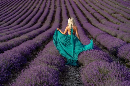 A woman with long blonde hair in a light green dress is walking through a lavender field alone waving a lavender dress