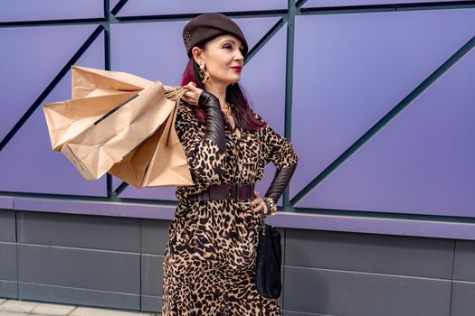 A happy shopaholic girl keeps her bags near the shopping center. A woman near the store is happy with her purchases, holding bags. Dressed in a leopard print dress. Consumer concept