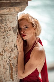Outdoor portrait of a young beautiful natural redhead girl with freckles, long curly hair, in a red dress, posing against the background of the sea