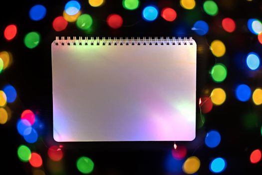 White notepad with defocused bokeh lights on black background, an abstract naturally blurred backdrop for Christmas eve or birthday party. Festive colorful lights in blur. Overlay effect for design