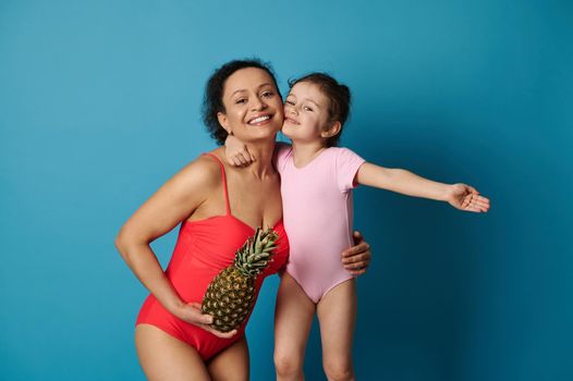 A cute little girl hugging her mother with a pineapple in her arms, both in bathing suits. Happy mothers day and summer concepts on blue background.