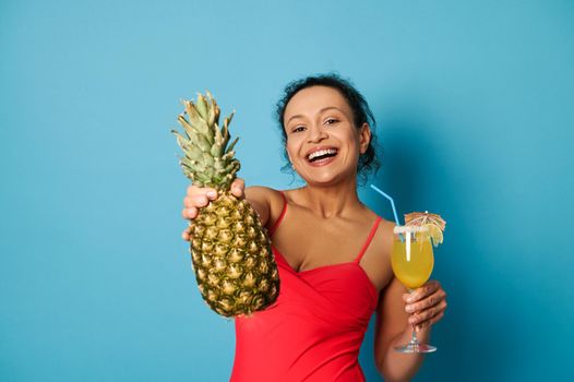 Charming brunette with beautiful smile holding a glass with cocktail and showing a pineapple to the camera. Summer concepts with space for text