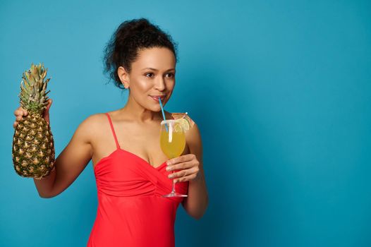 Young woman with a pineapple drinking a delicious exotic cocktail from a straw, posing over blue background with space for text. Summer concepts