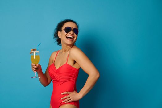 Happy woman in sunglasses and red bathing suit holding cocktail and smiling, posing on blue background with space for text