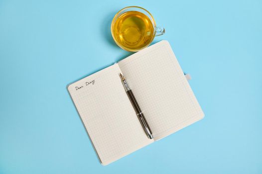 Open notepad with word Dear Diary and an ink pen next to a tea cup on blue background with copy space. High angle view, flat lay.