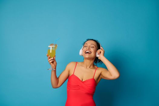 Happy brunette with closed eyes holding cocktail and enjoying listening to music with headphones isolated on blue background with copy space. Summer concept
