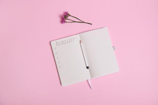 Still life. Open agenda, diary, notebook with a list to do on white sheet of paper in line with copy space, pencil in the middle of the agenda and meadow flower on pink background with space for text