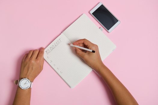 Flat lay of female hands holding pencil, writing in notebook, checking to-do list. Mobile phone lying on pink background with space for text. Business, planning and time management concept