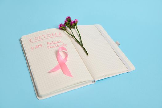 Flowers and pink ribbon, symbol of Breast Cancer Awareness Day, lying on a page with a line of notepad with inscriptions reminding of a medical check-up . October 1st, International Breast Cancer Day