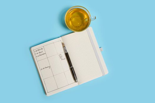 Flat lay composition of an open agenda with schedule, ink pen and transparent glass cup of tea lie on blue surface. Color background with copy space . Time management, deadline, schedule concept