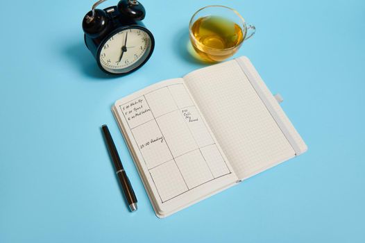 Time management, deadline and schedule concept: alarm clock on schedule plan, organizer with plans, ink pen and transparent glass cup with tea on blue background with space for text. Flat lay.