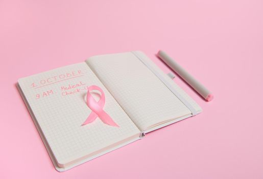 High angle view of a pink ribbon, on opened diary with inscriptions reminding of a medical check-up. October 1st, World Breast Cancer Day, Women's health care and medical concepts.