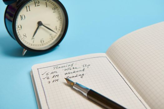 Cropped image of an open organizer with plans for the day, ink pen and alarm clock on blue background with copy space. Time management, business and planning concept