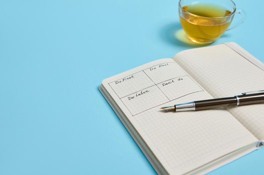 Time management, deadline concept: Cropped image of an open organizer notebook with timetable of the day by hour, ink pen, cup of tea on color background, copy space.
