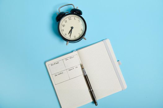 Flat lay of open notebook with timetable of the day by hour, pen, alarm clock on colored background with copy space. Time management, deadline and concept of proper planning and organization of time.