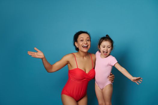 Mom and daughter in swimsuits smiling while posing over blue background with space for text