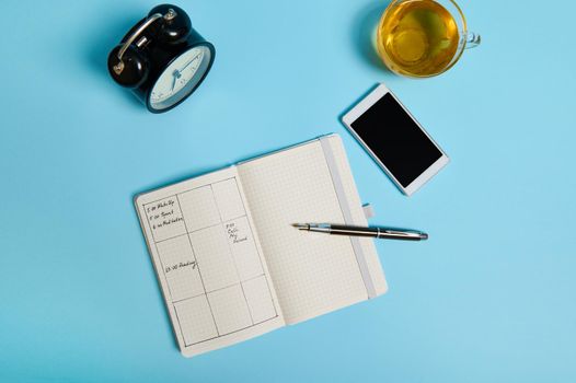 Flat lay of open agenda with schedule, smartphone, ink pen, glass cup of tea and black alarm clock lie on blue surface. Color background with copy space . Time management, deadline, schedule concept