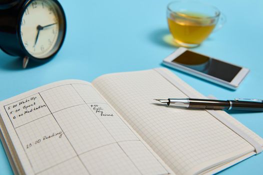 An open organizer with schedule, smartphone, ink pen, glass cup of tea and black alarm clock lie on blue surface. Color background with copy space . Time management, deadline, schedule concept