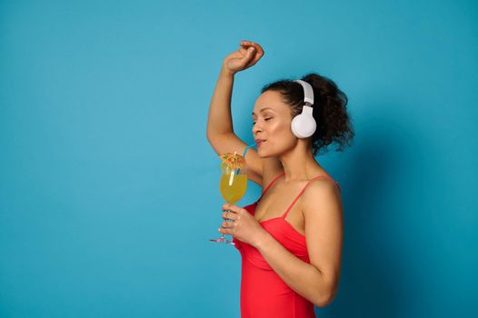 Side portrait of a woman in a red swimsuit drinking juice from a straw and enjoying listening to music with headphones. Isolated on blue background with copy space