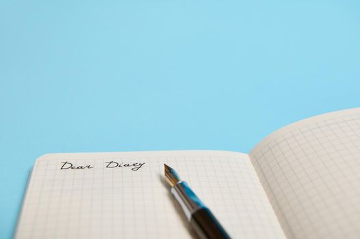 Cropped studio shot of an open notebook, organizer, diary with word Dear Diary and an ink pen isolated on blue background with copy space