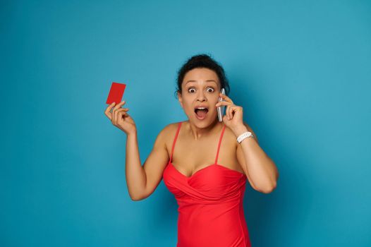 Surprised woman in red swimsuit talks on mobile phone and poses with open mouth and a blank plastic red card in her hand. Copy space