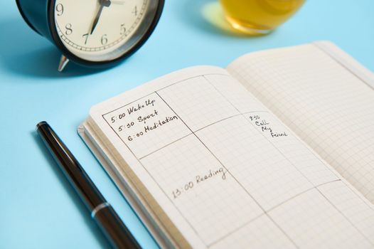 Cropped image of an open notebook with schedule, ink pen, black alarm clock lie on blue surface. Color background with copy space for text . Time management, deadline and schedule concept