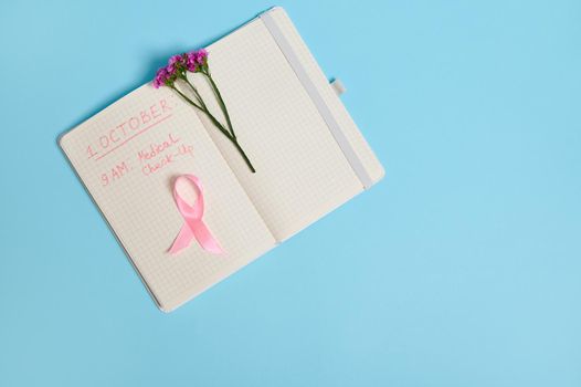 Flat lay with copy space of flower and pink ribbon, on opened notepad with inscriptions reminding of a medical check-up. October 1st, World Breast Cancer Day, Women's health care and medical concept.