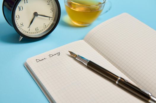 Cropped shot of an open notepad with word Dear Diary, alarm clock and an ink pen next to a tea cup on blue background with copy space