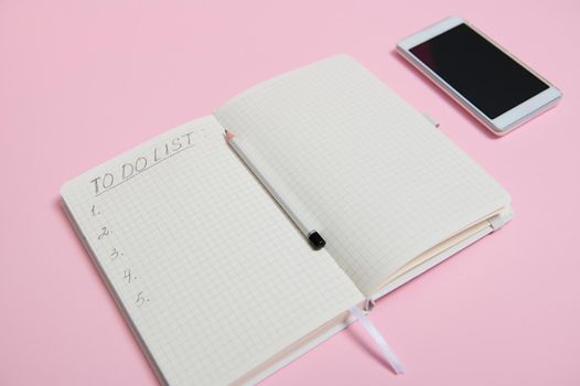 Still life. A white mobile phone lying down on pink background next to a pencil on the middle of an open agenda, diary, notebook with a list to do on white sheet of paper in line with copy space