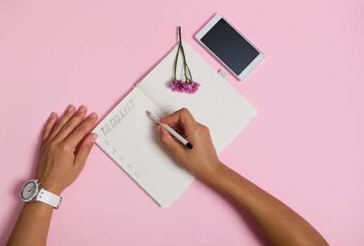 Flat lay of female hands holding pencil, writing in notebook, checking to-do list. Mobile phone and pink flower lying on pink background with copy space. Business, planning and time management concept