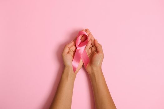 Top view of hands holding pink ribbon on pink background, Breast cancer awareness, October Pink day, World Wancer Way, national Cancer Survivor Say. Women's health care and medical concept. Copy space