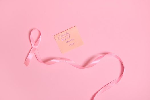 High angle view of a long pink ribbon with endless one end, and a paper note with lettering 1 October Breast Cancer Awareness , isolated on pink background with copy space. Flat lay