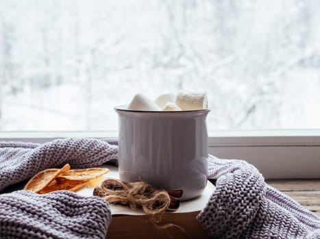 Relaxing winter day at home with traditional winter hot drink. Coffee with marshmallows, cinnamon, book and a cozy grey sweater on vintage windowsill against snow landscape from outside. Soft focus