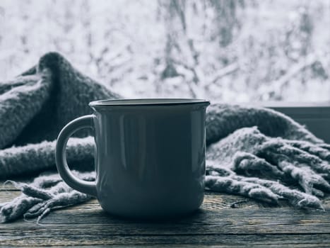 Scandinavian hygge style. Mood, lifestyle, still life concept. Hot cup of coffee and cozy grey scarf on vintage windowsill against snow landscape from outside. Relaxing winter day at home. Soft focus