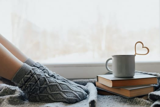 Cozy winter still life: woman legs in warm woolen socks under shaggy blanket and mug of hot beverage on old windowsill against snow landscape from outside.