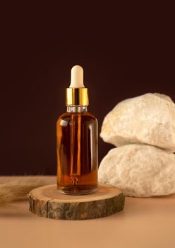 Transparent glass cosmetic dropper standing on wooden podium near stones and dry flowers on dark beige background. Beauty composition. Concept of natural and original skincare products presentation.