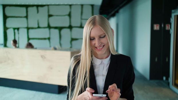 Pretty blond woman with long hair wearing elegant black suit and smiling charmingly at camera inside of modern office