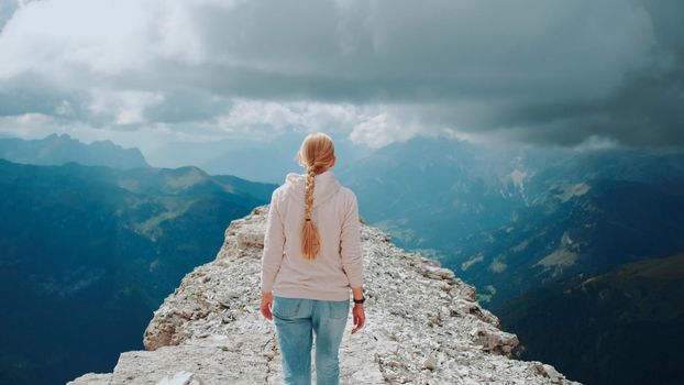 Back view of blonde woman walking on the top of the mountain under the clouds. Stunning view